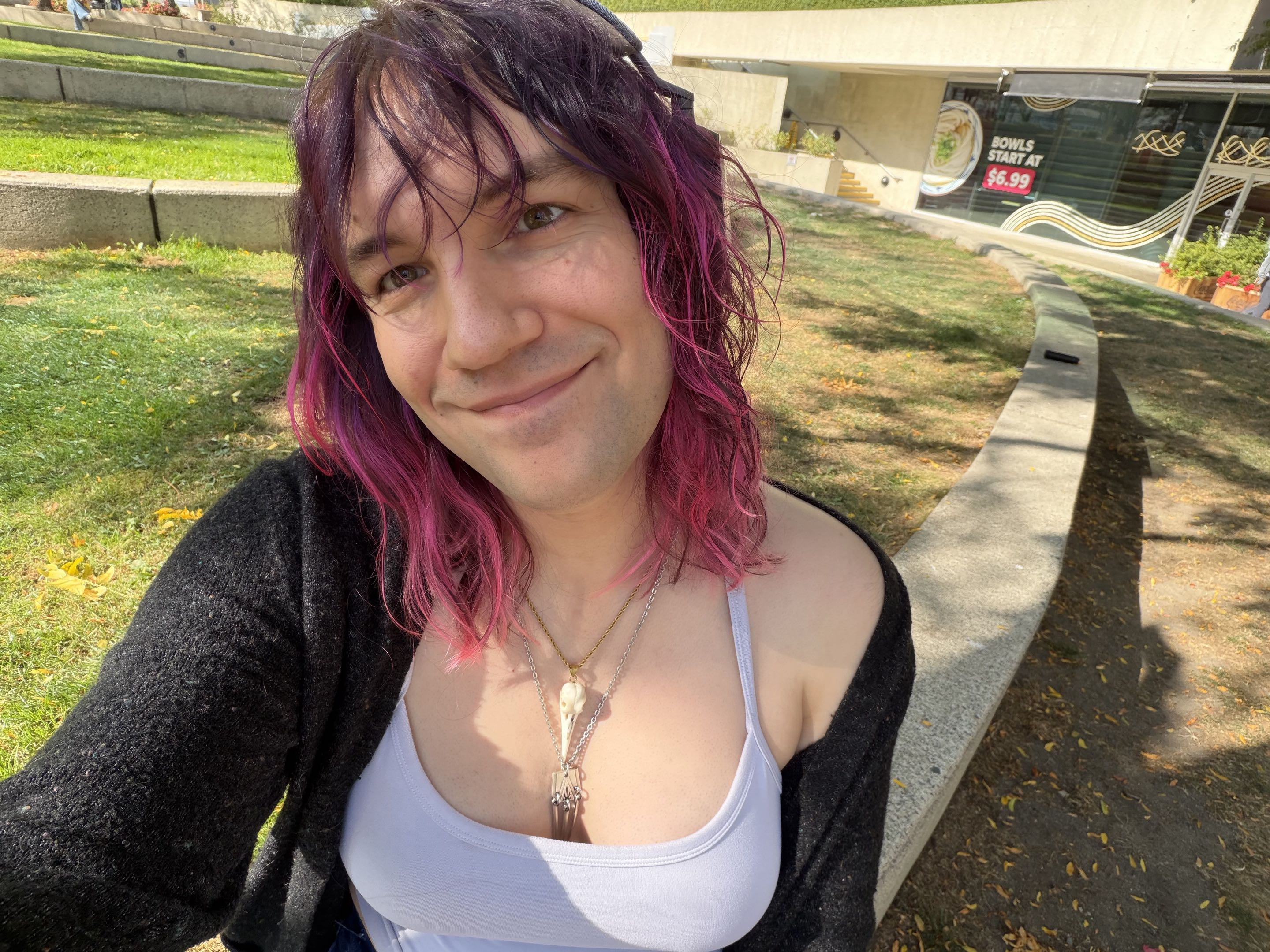 A selfie of me wearing a cardigan and cami top, sitting outside on some grass near a noodle restaurant